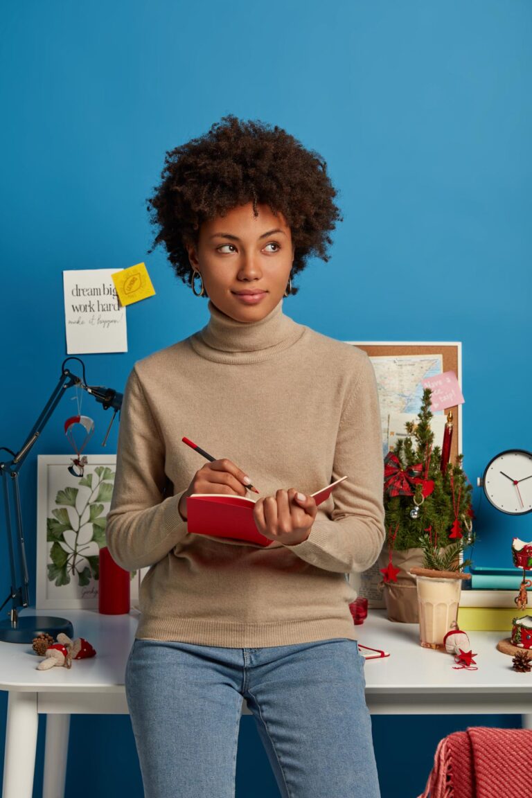 Indoor shot of contemplative Afro American woman holds diary, writes notes, thinks over plans and tasks to do, prepares for Christmas holiday, stands near desktop decorated with special attributes